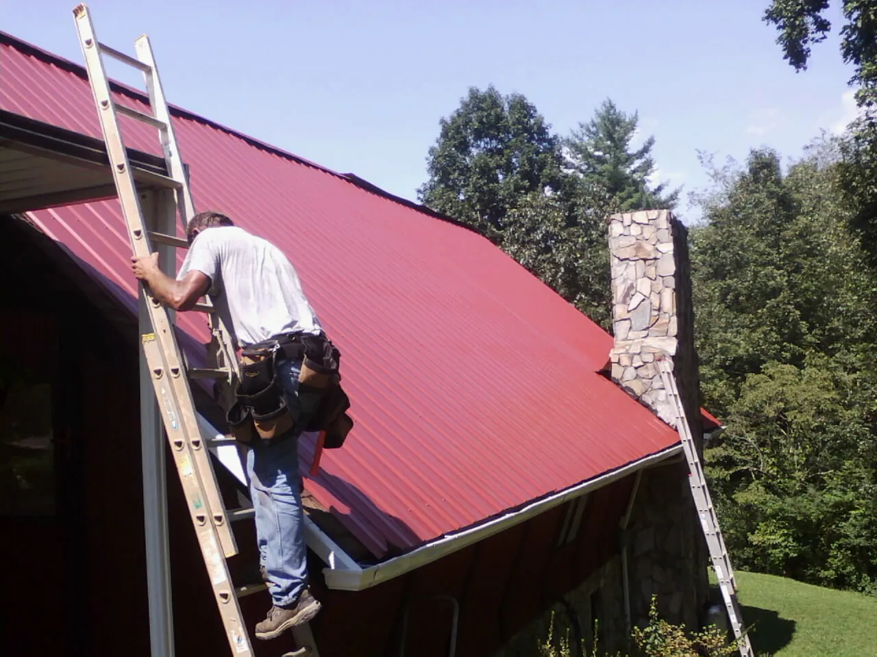 Roofer on a ladder beside a red metal roof and stone chimney — Soffit & Fascia Repair by Roofing Brothers in Hawley